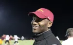 Smiling man in red cap and black jacket at nighttime sports field with players in background