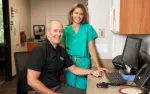 Smiling male doctor and female nurse in green scrubs at a medical office desk with computer and phone.
