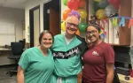 Three smiling medical staff celebrating a birthday with balloons and a banner in an office setting.