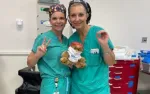 Two smiling female surgeons in teal scrubs holding a teddy bear inside a medical facility.