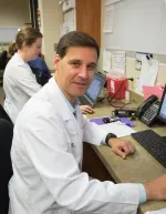 Male doctor in white coat working on computer in medical office with female colleague in background.