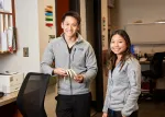 Two smiling people in gray fleece jackets standing in an office with books and a 3D spine model.