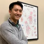 Smiling man in a gray jacket stands in front of an anatomical hand and wrist chart on a beige wall