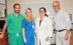 Group of four healthcare professionals smiling indoors wearing scrubs and a white coat in medical office.