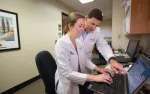 Two medical professionals in white coats working together on a laptop in a clinical office environment.