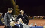 Two men attend to a young male athlete lying on a table at night on a running track during an outdoor sports event.