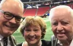 Three smiling older adults taking a selfie at an indoor stadium with a green field and red seats in the background