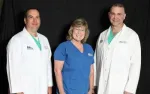 Three medical professionals smiling, two men in white coats and a woman in blue scrubs, posed against a black background