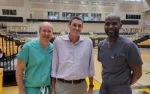 Three men, two in medical scrubs and one in casual shirt, posing inside a sports arena with empty stands.