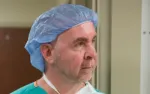Middle-aged male healthcare worker wearing a blue surgical cap and green scrubs looking to the side indoors.