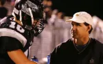 Football player in black helmet listens to coach wearing white cap during nighttime game.