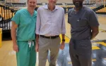 Three men standing on a gymnasium floor, two in medical scrubs and one in casual business attire, smiling at camera