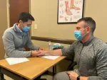 Doctor wearing a mask examining patient's wrist in a medical office with anatomy chart on the wall