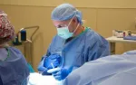 Surgeon in blue scrubs and mask performing a medical procedure in an operating room with surgical tools.