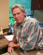 Mature man in plaid shirt sitting at desk with laptop and documents in home office with window background