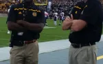 Two football coaches stand with arms crossed on the sidelines of a green football field during a game.