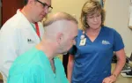 Two medical professionals discuss a treatment plan with a bald patient in green scrubs inside a clinic room.