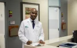 Confident male doctor in white coat writing on clipboard at hospital reception desk.