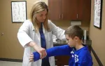 Female doctor examines young boy's arm in medical office with anatomical charts and brown cabinets