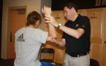 Man in black shirt demonstrating arm technique to another man in gray Adidas shirt in a sports locker room