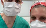 Two healthcare professionals wearing masks and scrubs in a clinical setting with safety glasses and head covering.