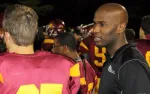 Football coach talks to players wearing maroon and yellow uniforms during a nighttime game.
