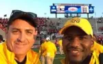 Two smiling men in yellow jackets and hats at a sports stadium with a crowd and scoreboard in the background