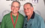 Two older men smiling, one in a green shirt with a red lanyard, the other in a blue shirt, against a branded backdrop