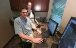 Man and woman working together at office desk with computer monitors and window view of street outside