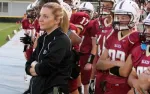Female football coach standing with arms crossed as players in maroon uniforms and helmets stand behind her on sidelines.