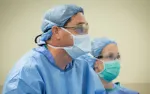 Two medical professionals wearing surgical masks, caps, and gowns in a clinical setting focused on patient care.