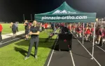 Man in black stands with arms crossed near pinnacle-ortho.com tent at nighttime football field event with crowd and cheerleaders.