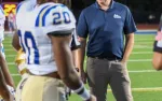 Football coach in navy polo and khaki pants standing on field with players in white and blue uniforms at night.