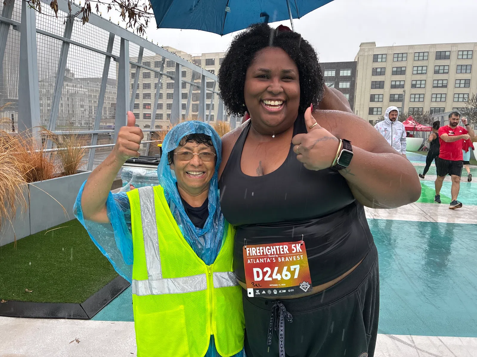 Two smiling women giving thumbs up at a rainy outdoor firefighter 5K event in urban Atlanta surroundings.