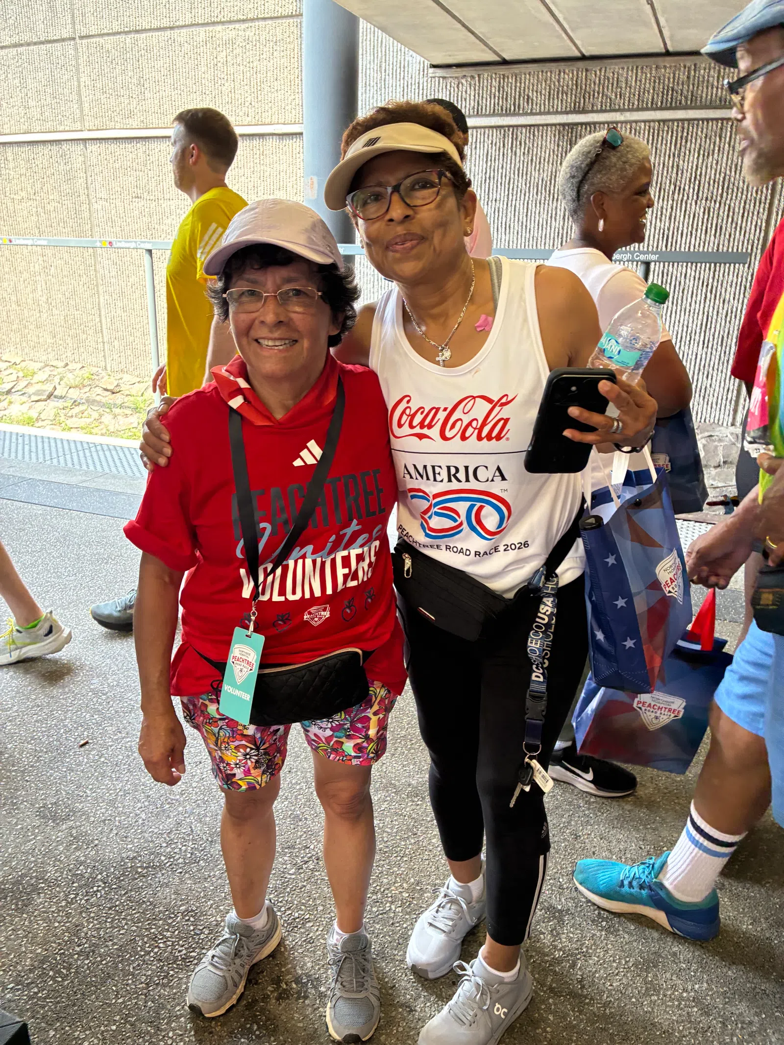 Two smiling women at a community event wearing volunteer and Coca-Cola race shirts outdoors