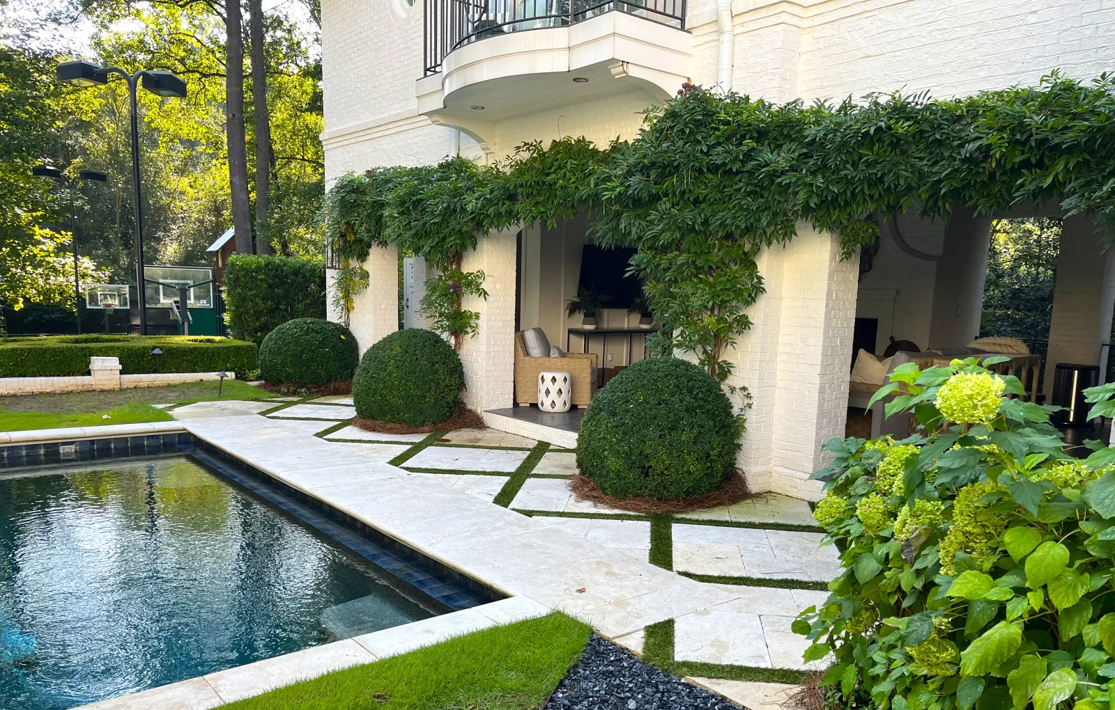 Luxury Atlanta backyard with custom outdoor living space featuring poolside lounge area, manicured boxwood shrubs, climbing greenery on pergola, and elegant stone pavers with grass joints designed by Gibbs Landscape Company.