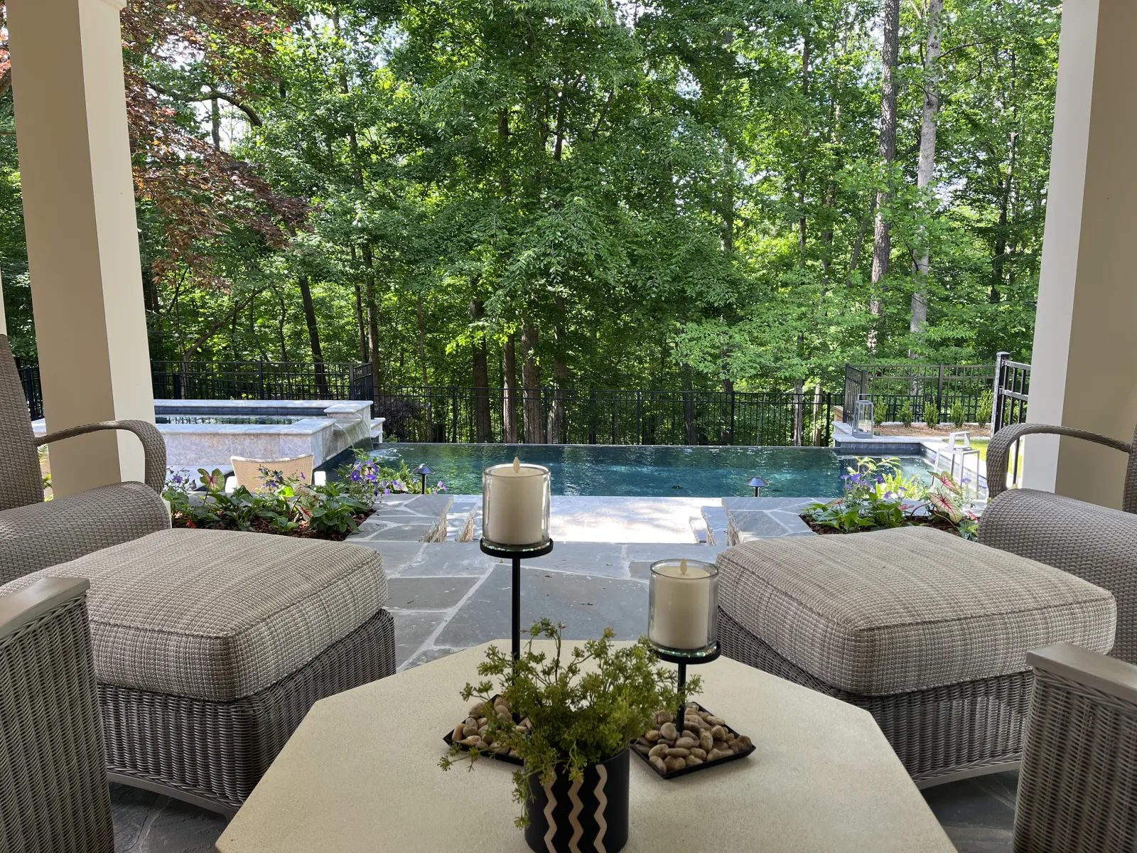Luxury covered patio in Atlanta overlooking an infinity-edge pool with forest view, featuring wicker lounge furniture, elegant stone flooring, potted plants, and tranquil outdoor ambiance, designed by Gibbs Landscape Company.
