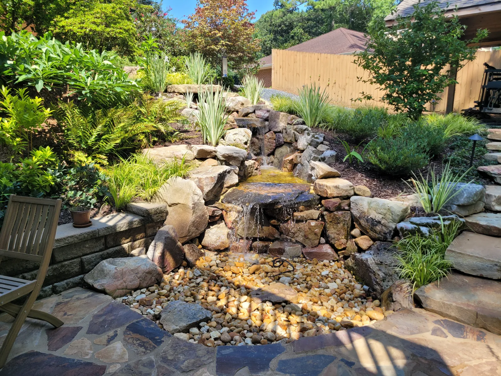 Serene backyard water feature in Atlanta with cascading stone waterfall, natural rock edging, lush plantings, and flagstone patio seating area, custom designed by Gibbs Landscape Company.