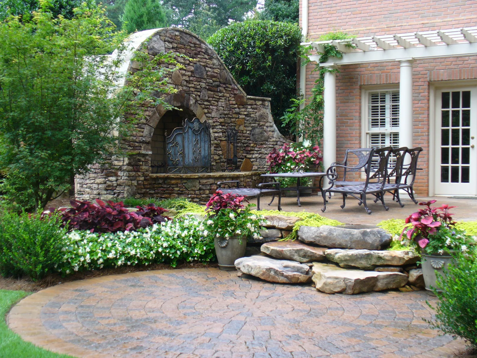Elegant Atlanta backyard patio with rustic stone fireplace, wrought iron seating, lush seasonal flower beds, natural stone steps, and pergola-covered brick exterior, professionally designed by Gibbs Landscape Company.