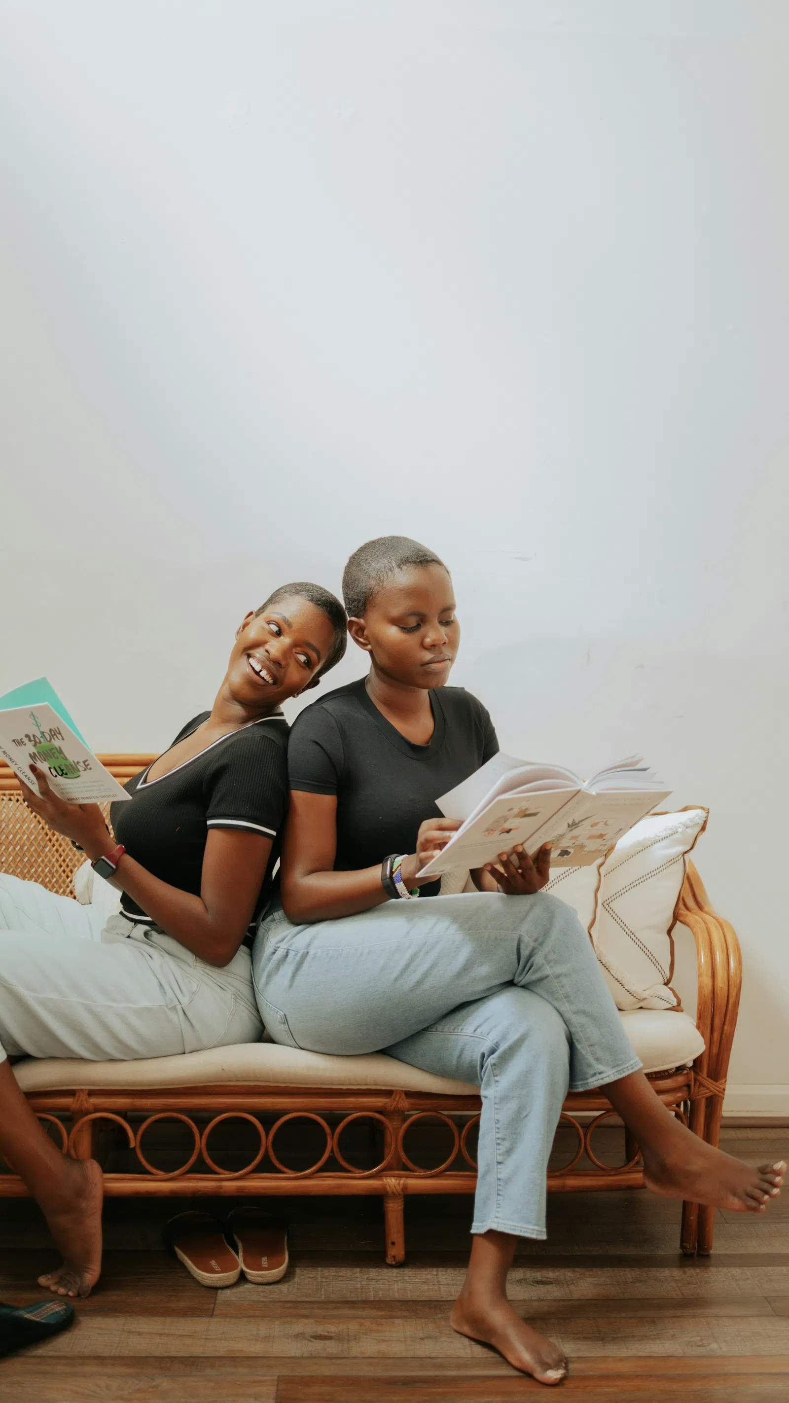 Two women sitting back-to-back on a wicker sofa reading books, relaxed and barefoot indoors.