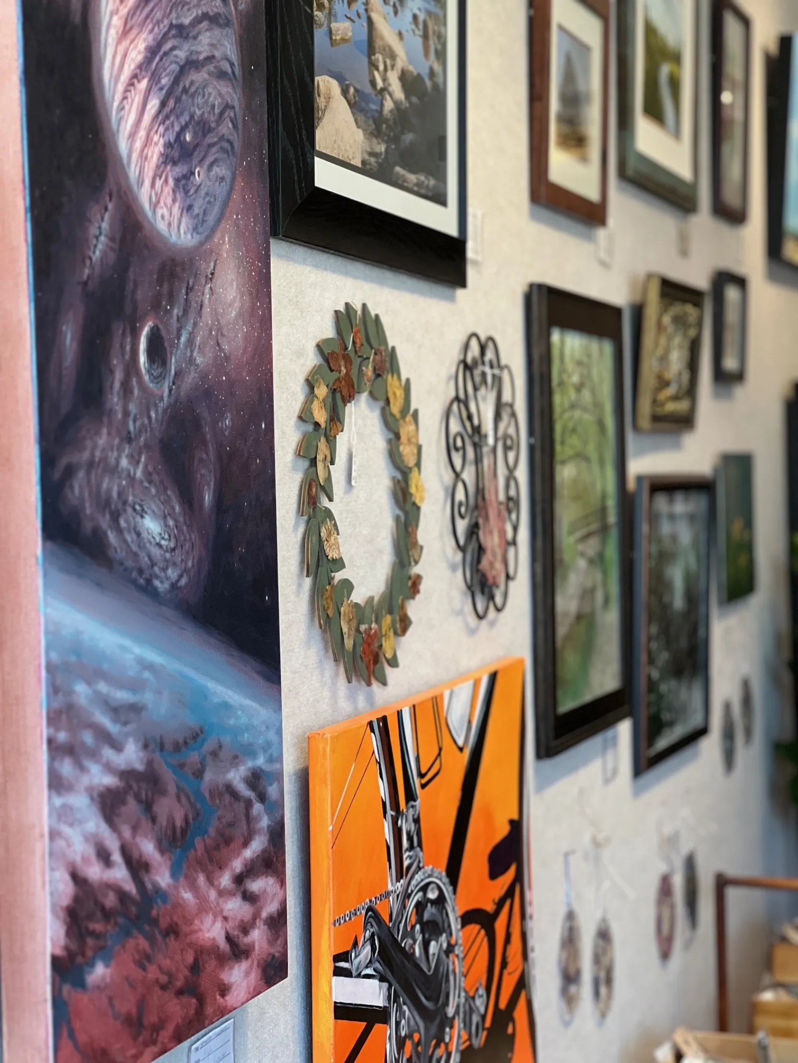 Wall display of various framed art pieces including a space scene, a bicycle painting, and decorative wreaths in a gallery.