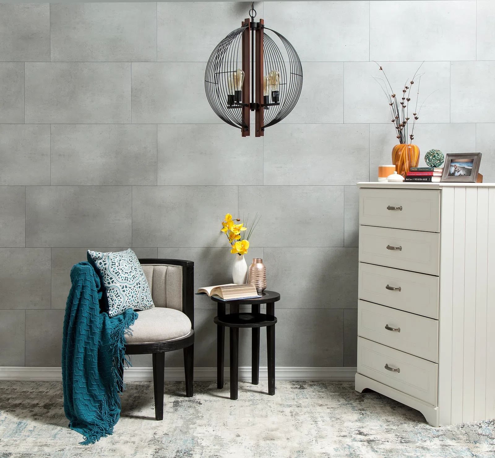Cozy reading corner with armchair, teal throw, side tables, flowers, and white dresser against gray wall.