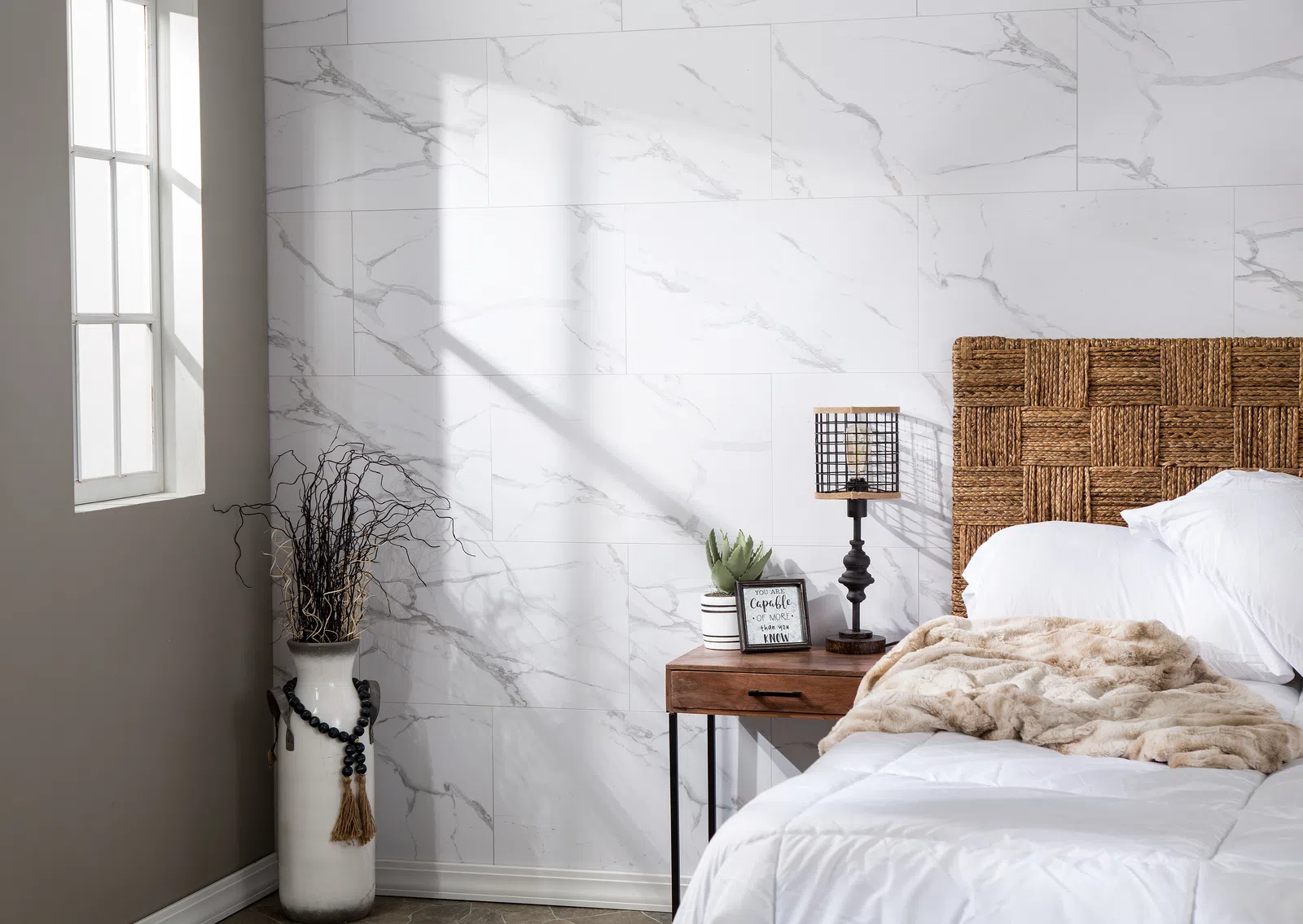 Bright bedroom corner with wicker headboard, white bedding, wooden nightstand, lamp, and decorative vase by window