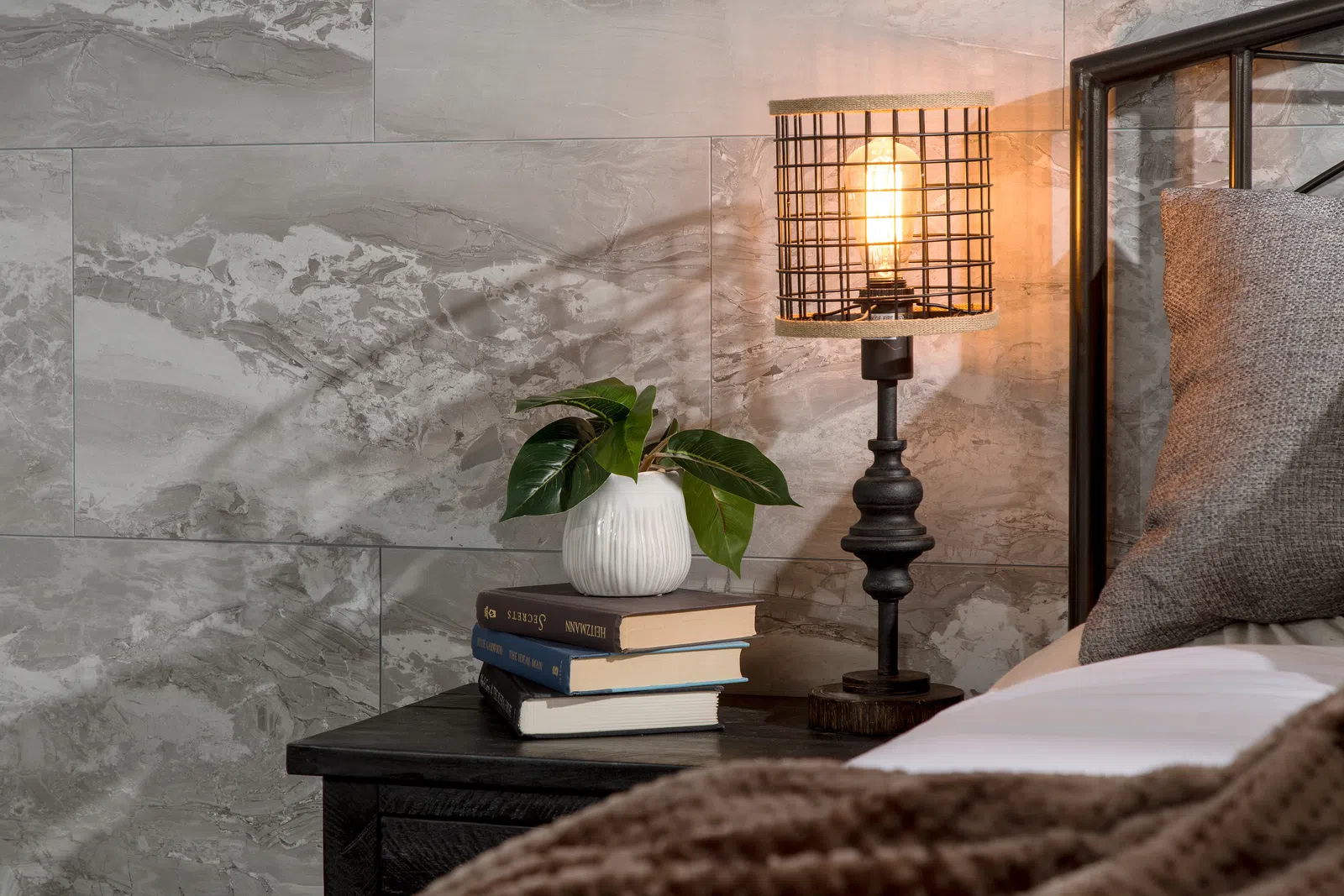 Cozy bedroom scene with a nightstand holding books, a white vase with green leaves, and a lit rustic lamp beside a bed.