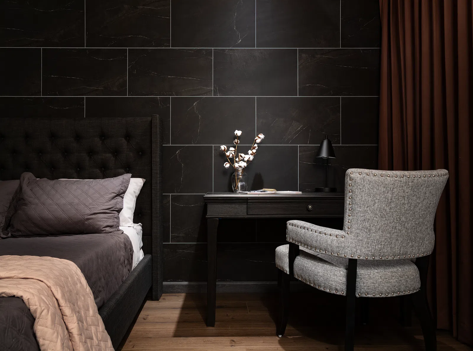Modern bedroom corner with dark tiled wall, upholstered bed, black desk, gray chair, and brown curtains.
