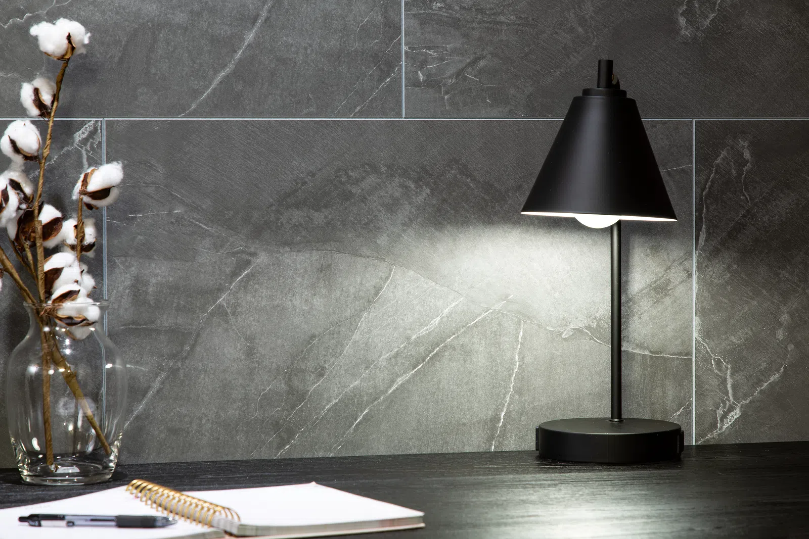 Modern black desk lamp illuminating workspace with notepad and cotton stems in glass jar on dark surface.