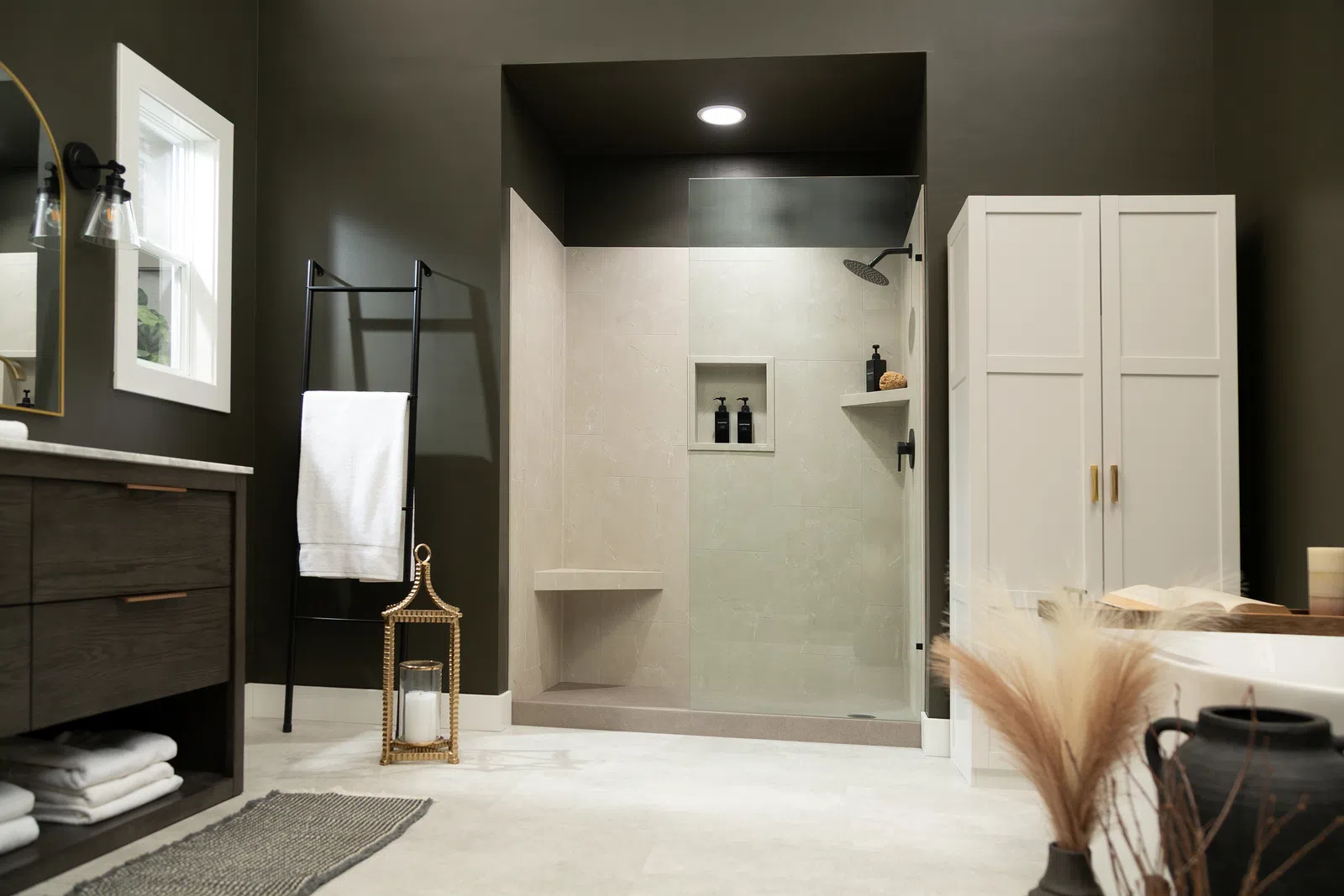 Modern bathroom with walk-in shower, dark walls, white towels, wooden vanity, and decorative lantern and vase.