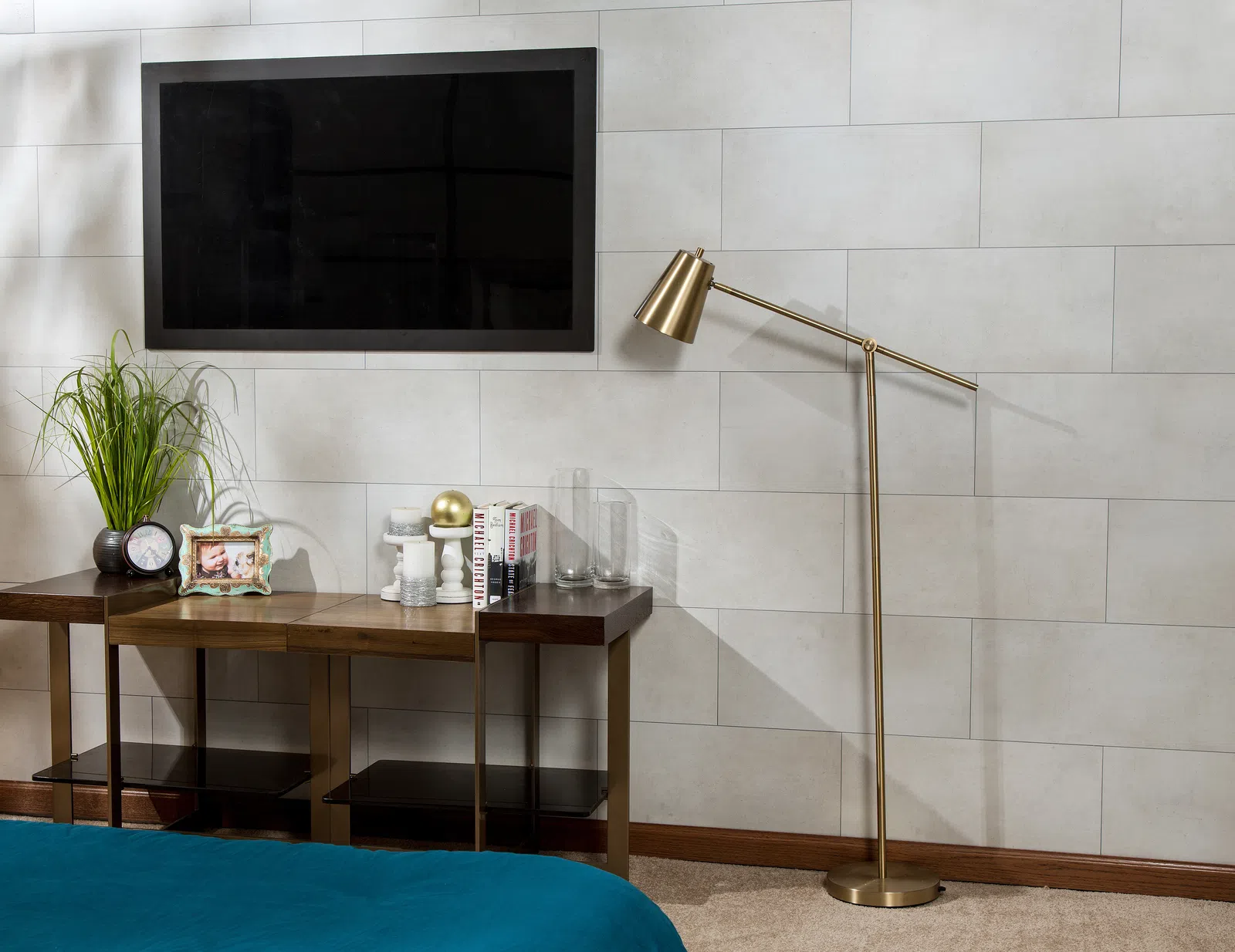 Modern bedroom corner with wall-mounted TV, wooden console table, gold floor lamp, and decorative plants and books.
