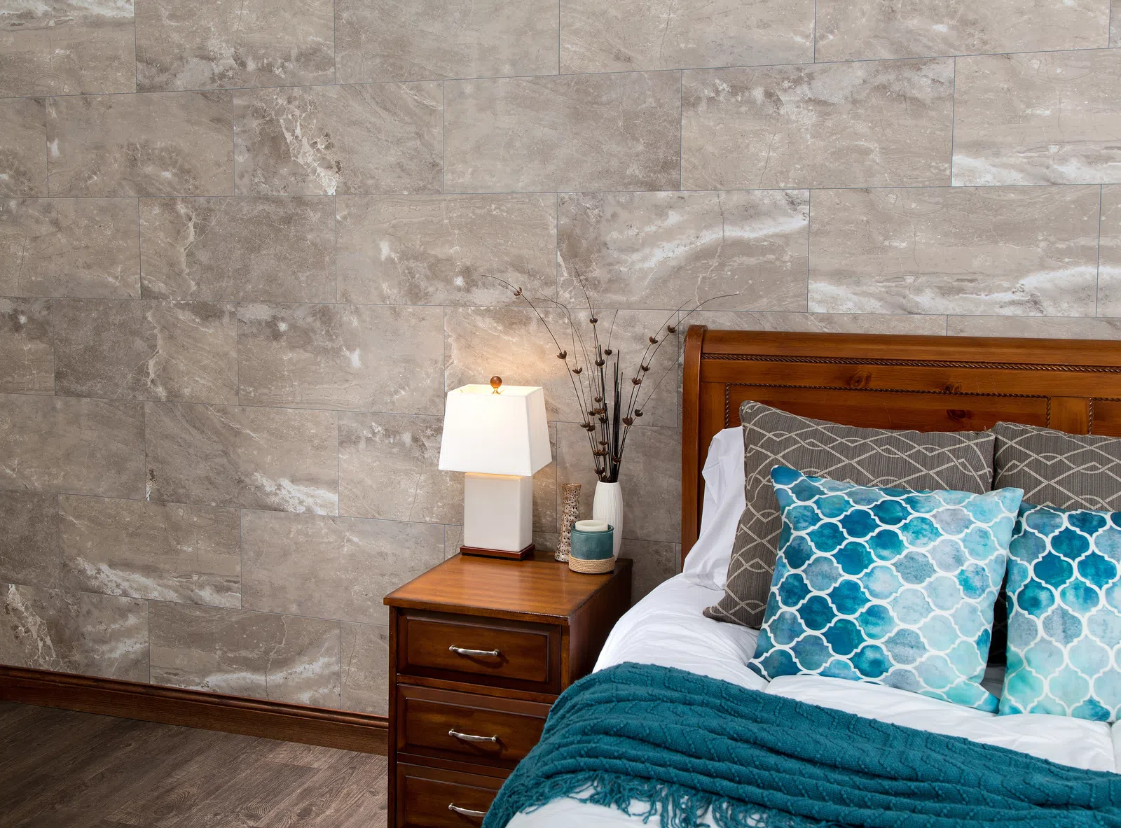 Cozy bedroom corner with wooden bed, blue patterned pillows, teal throw, nightstand, and table lamp on marble wall.