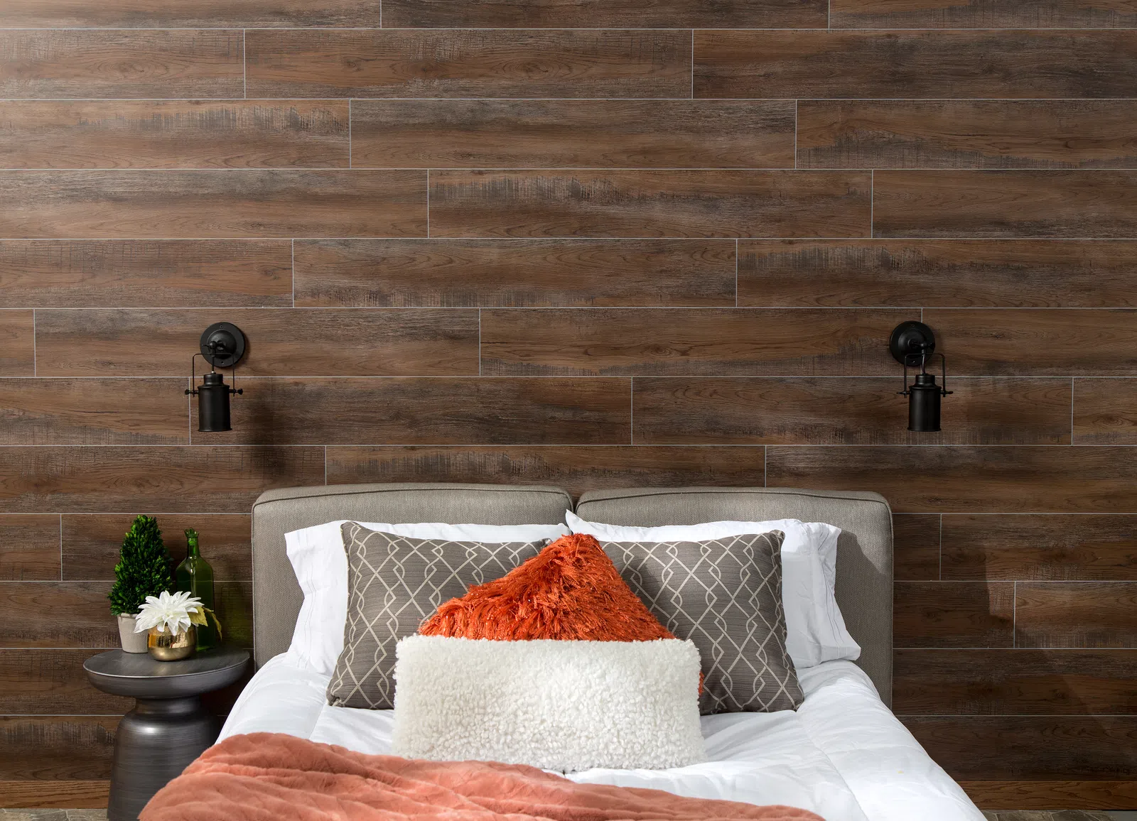 Cozy bed with textured pillows and throw against a dark wooden plank accent wall with wall-mounted lamps.
